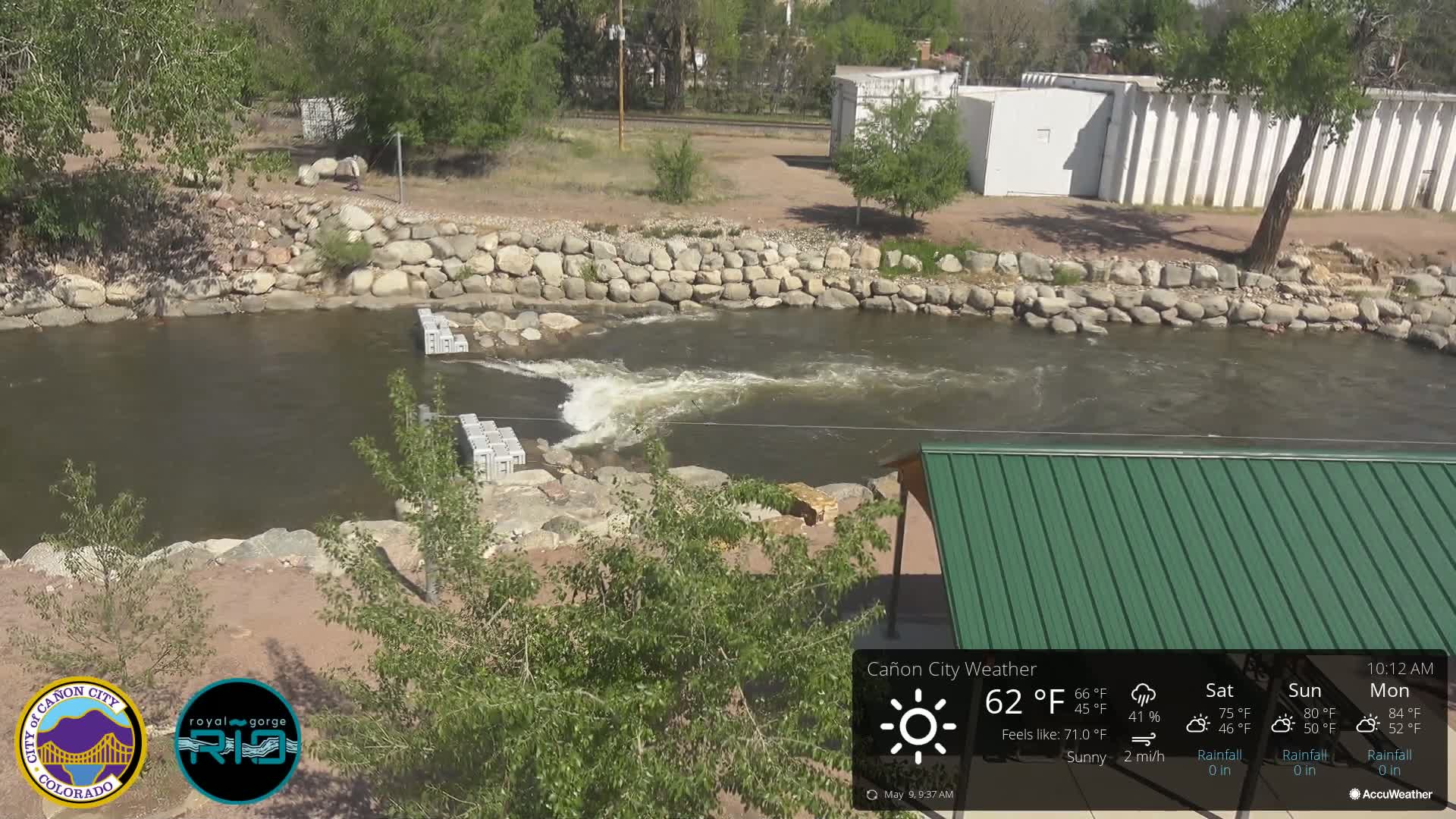 A river flows under a sunny sky past a rocky bank, a small dam, and a green roofed structure.