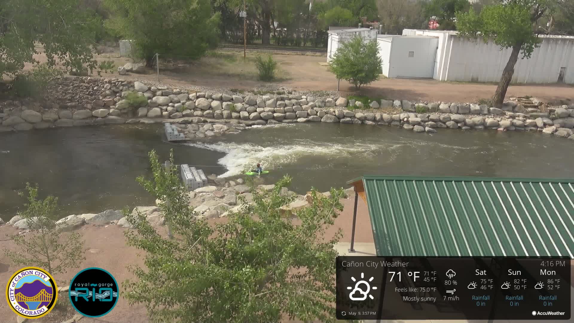 A person kayaks on a river under mostly sunny skies, with a rock wall and a green roofed structure visible.