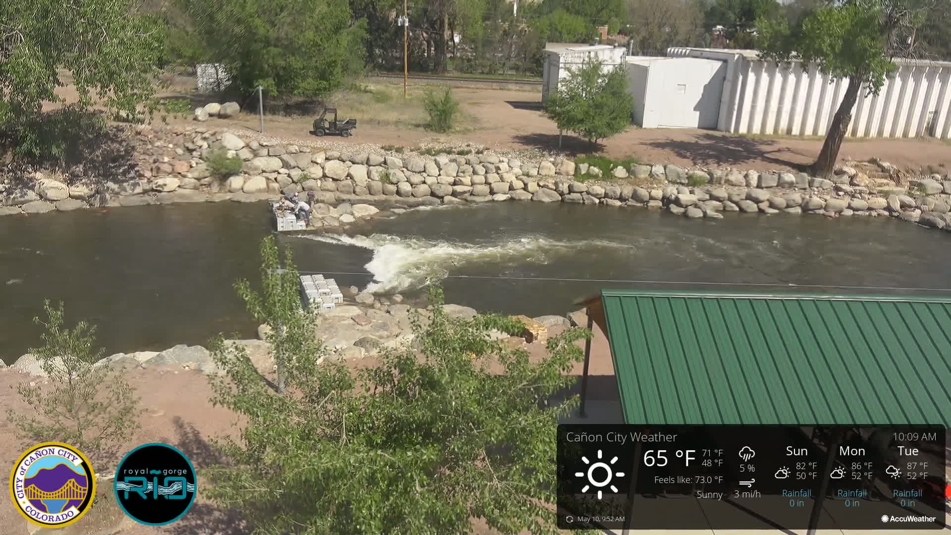 A sunny day shows people working near a river with a rocky bank and a green-roofed structure nearby.