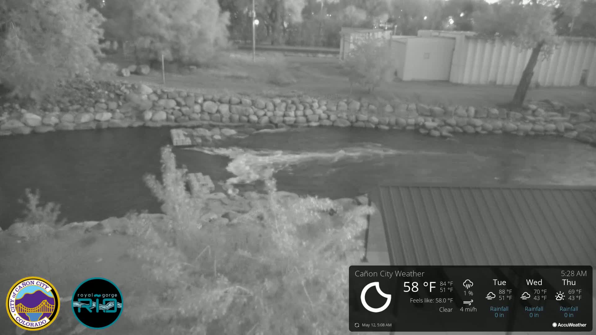 A nighttime grayscale image shows a river flowing under a clear sky, bordered by a low stone wall and vegetation.