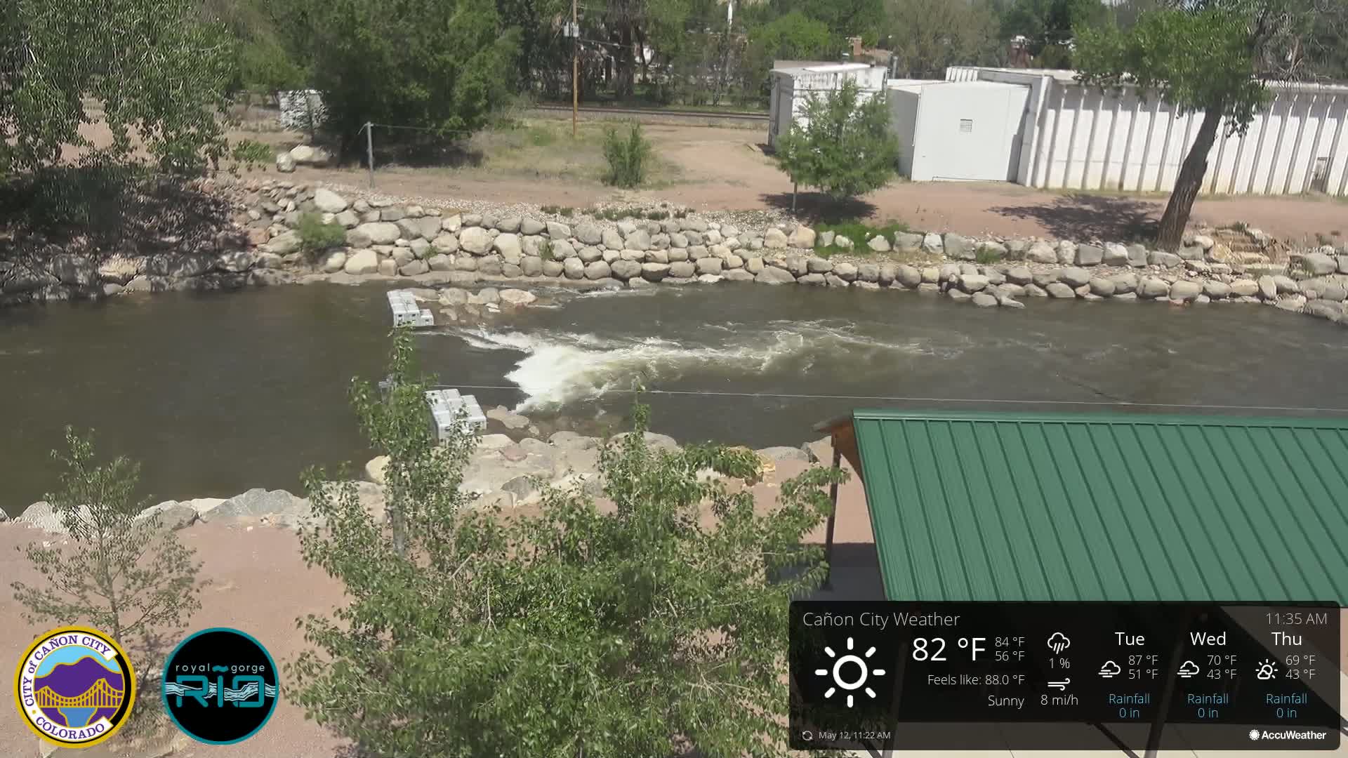 A rocky river flows under a sunny sky, past a low stone wall and a green roofed structure.