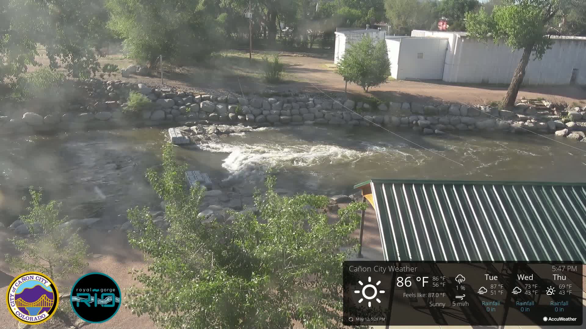 A rocky river flows under a sunny sky past a stone retaining wall and a partially visible structure with a green metal roof.
