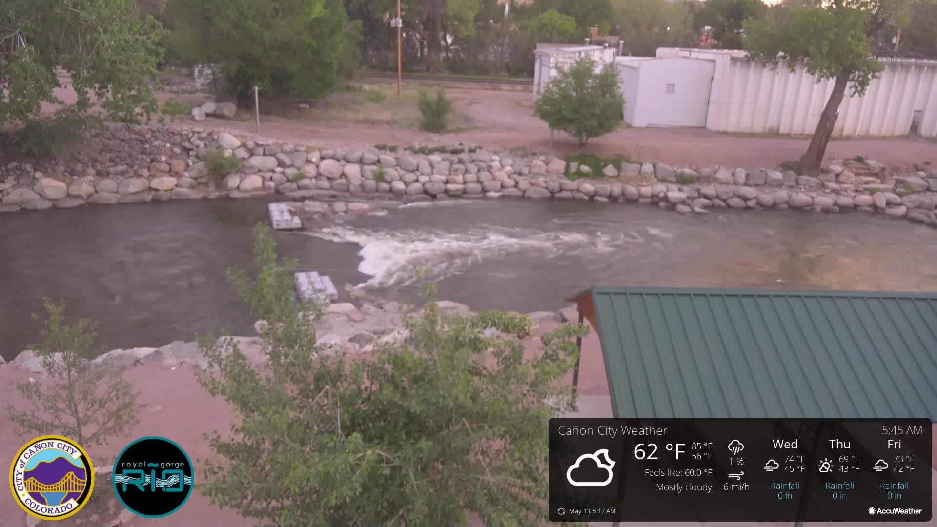 A mostly cloudy morning scene shows a river flowing over a low rock dam, with a green-roofed structure partially visible in the lower right corner.
