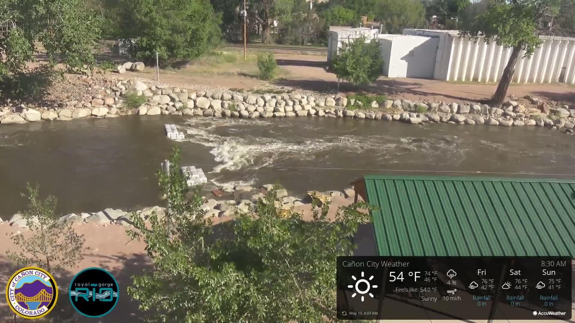 A swiftly flowing river, partially dammed with concrete blocks, runs alongside a rocky riverbank under sunny skies.