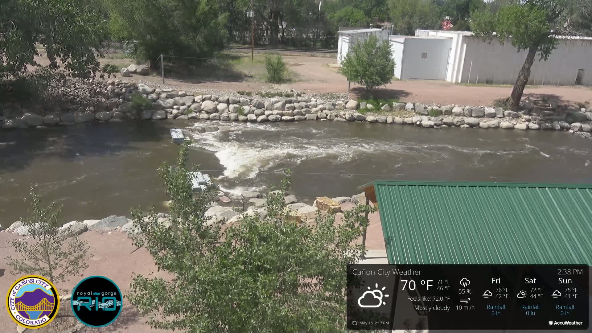 A mostly cloudy day shows a river flowing over a rocky section, with some buildings and trees visible in the background.