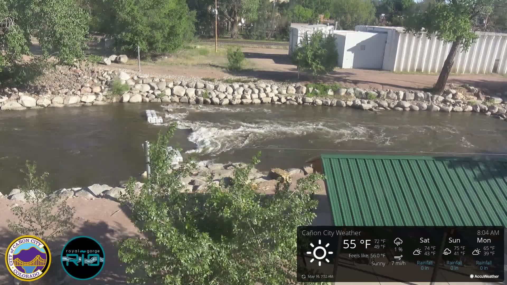 A rocky riverbank with a swiftly flowing river under sunny skies is partially visible beneath a green metal roof.