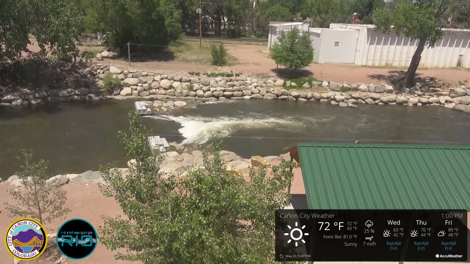 A river flows under a sunny sky past a rock retaining wall and a green roofed structure.