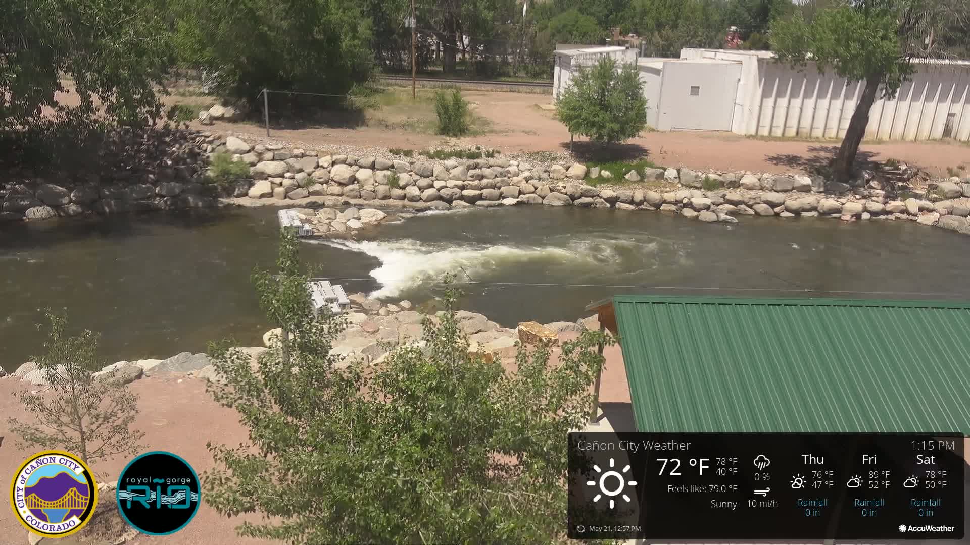 A sunny day shows a river flowing over a low dam, surrounded by rock walls and green vegetation; a green metal roof is partially visible in the foreground.