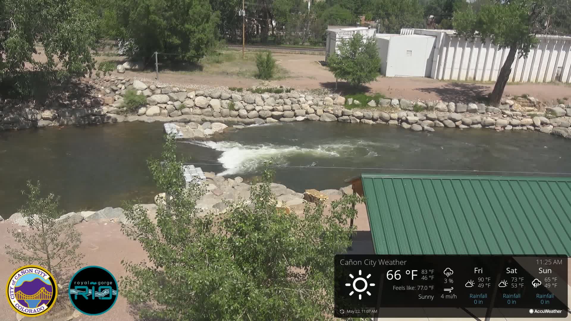 A rocky river flows under a sunny sky, cascading over a low dam built of large stones.
