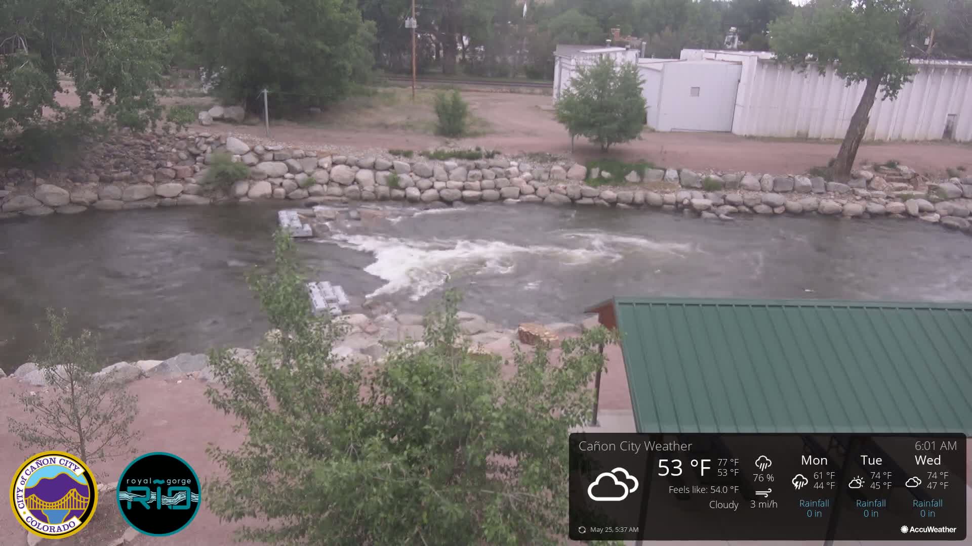 A cloudy day shows a river flowing over a low dam, with a rocky riverbank and green vegetation alongside a green roofed structure.