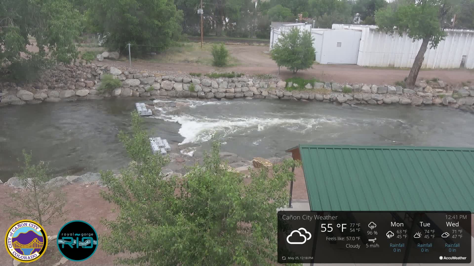 A cloudy day shows a river flowing over rocks and through a man-made stone dam, partially obscured by vegetation and a green roofed structure.