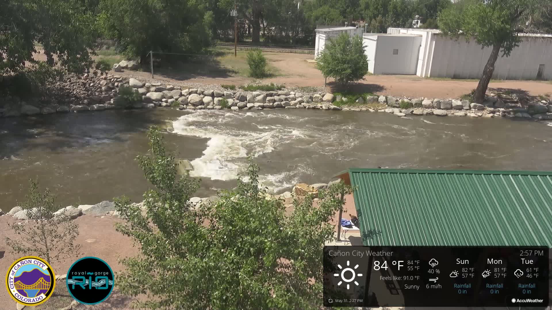 A rocky river flows under a sunny sky past a green-roofed structure and some buildings.
