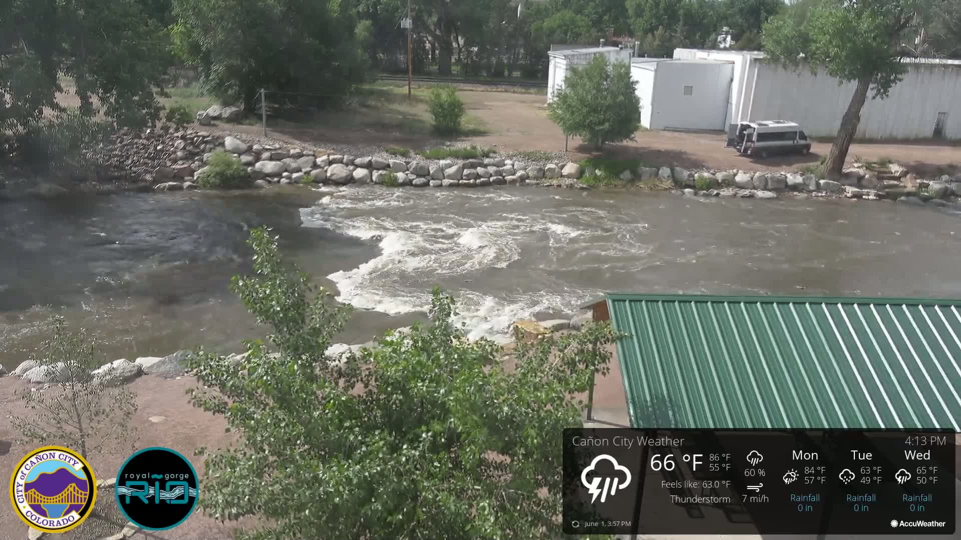 A fast-flowing river with whitewater rapids is seen under a thunderstorm, near a building and some trees.