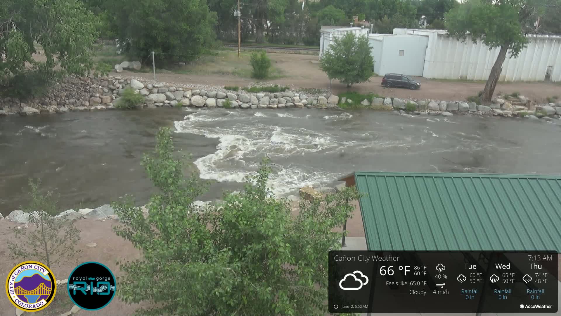 A fast-flowing river with some whitewater rapids is seen from a high vantage point on a cloudy day, next to a stone riverbank and a green roofed structure.