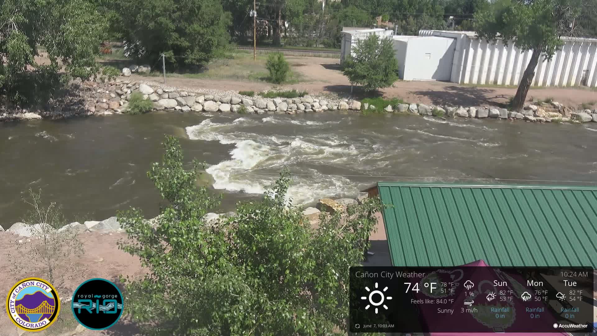A rocky riverbank with a swiftly flowing river under sunny skies is partially visible from behind leafy green trees and a green metal-roofed structure.