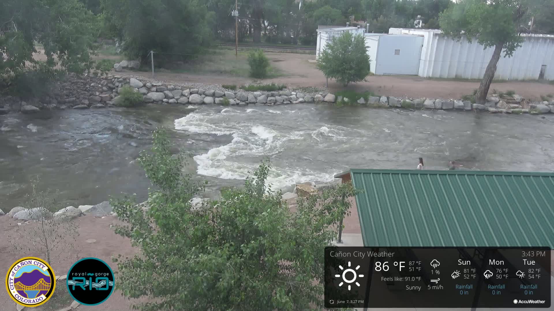 A swiftly flowing river, under sunny skies, passes by a rock embankment and a partially visible green roofed structure.
