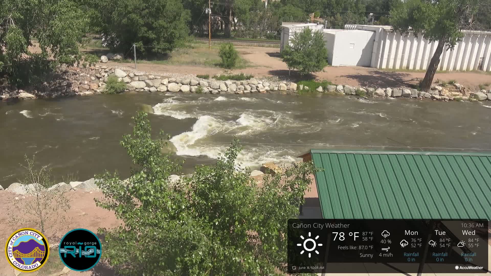 A fast-moving river with small waves flows past a rocky bank and a green-roofed structure under sunny skies.