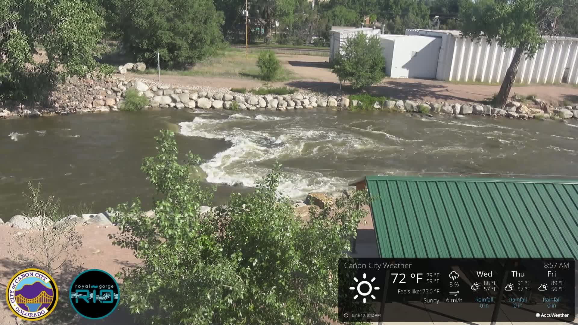 A swiftly flowing river with moderate rapids is seen under sunny skies, bordered by rock retaining walls and lush green vegetation, partially obscuring a green metal roofed structure.