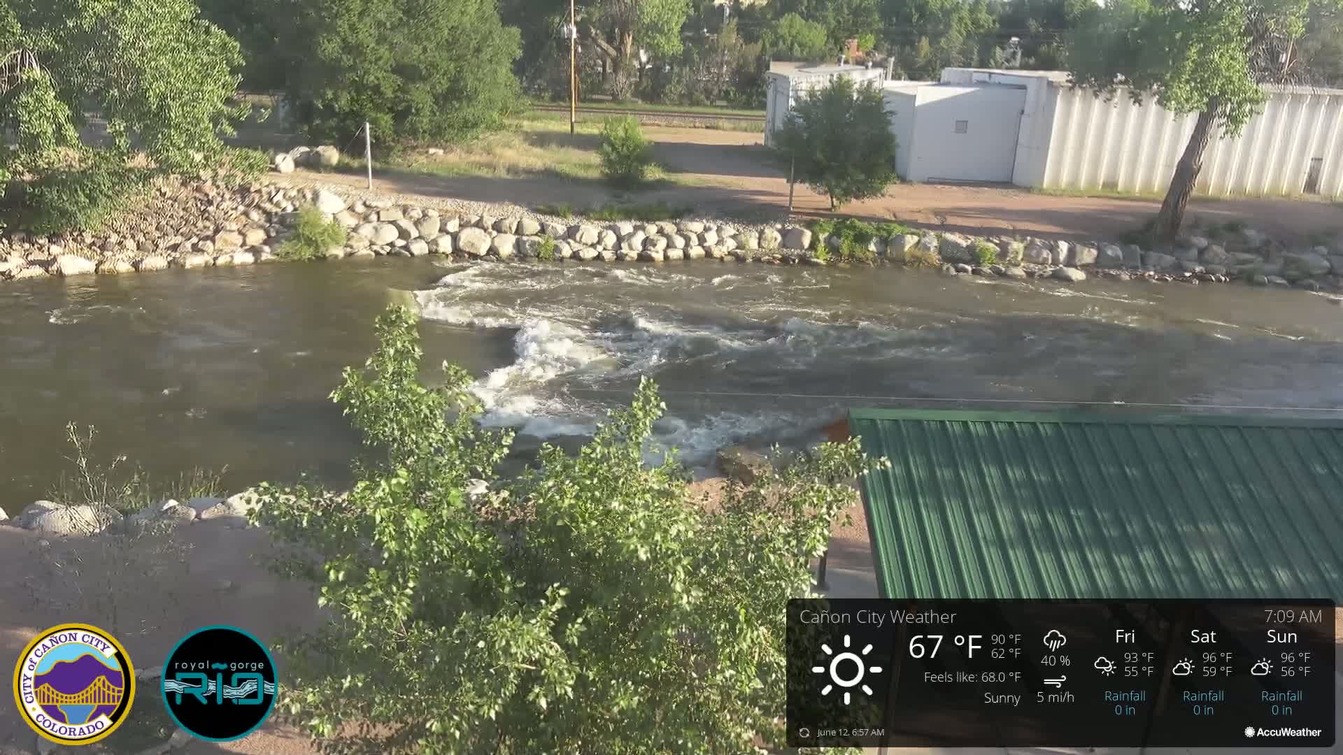 A swiftly flowing river with small rapids is partially visible under a sunny sky; the riverbank is lined with rocks and green vegetation, partially obscuring a building's green roof.