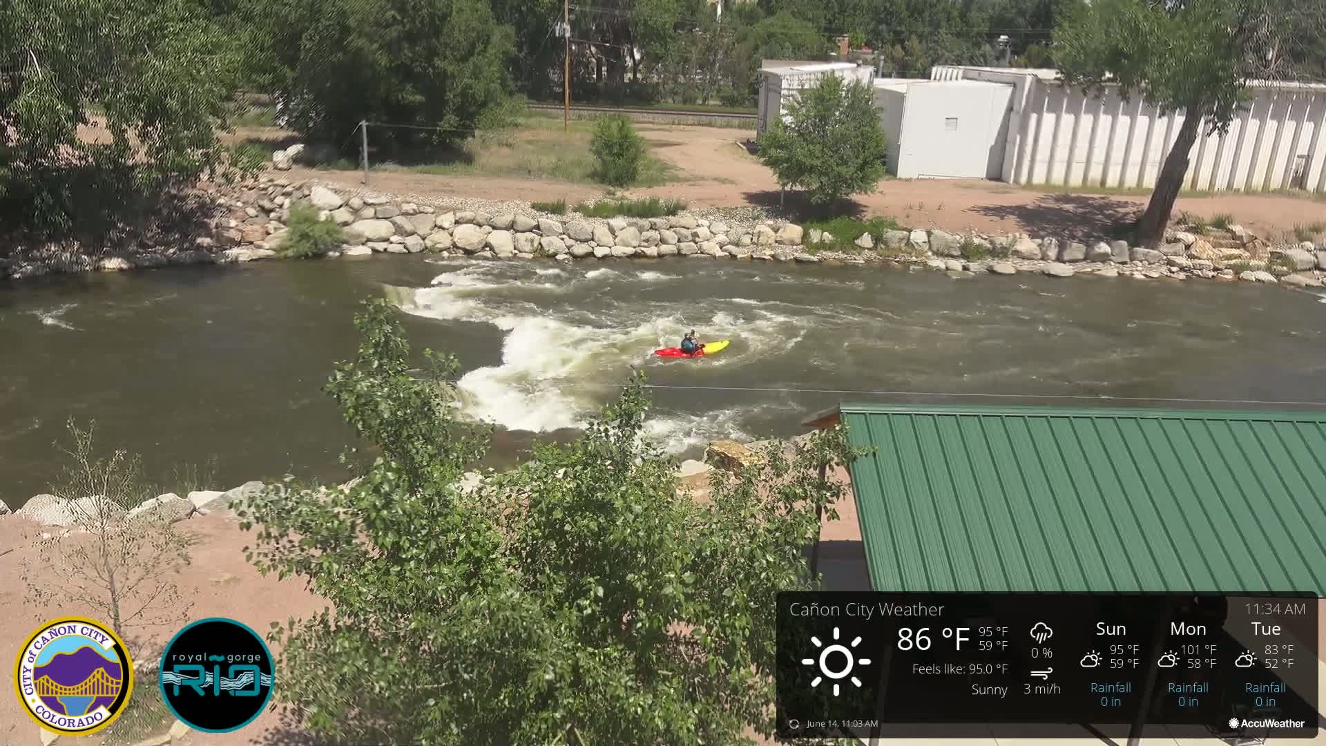 A person kayaks on a river under sunny skies, with a green-roofed structure visible in the foreground.