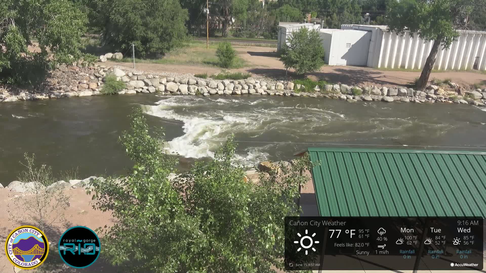 A fast-moving river flows under a sunny sky past a rock-lined bank and a green metal shelter.