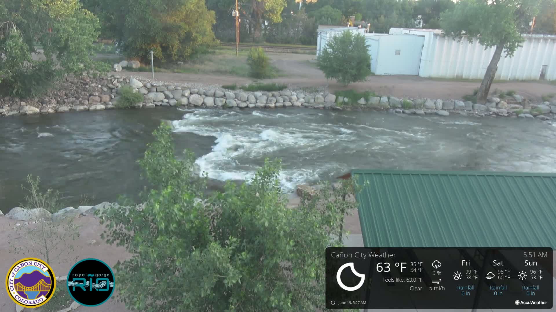 A swiftly flowing river, partially bordered by rock retaining walls, is seen under a clear sky; a portion of a green metal roof is visible in the bottom right.