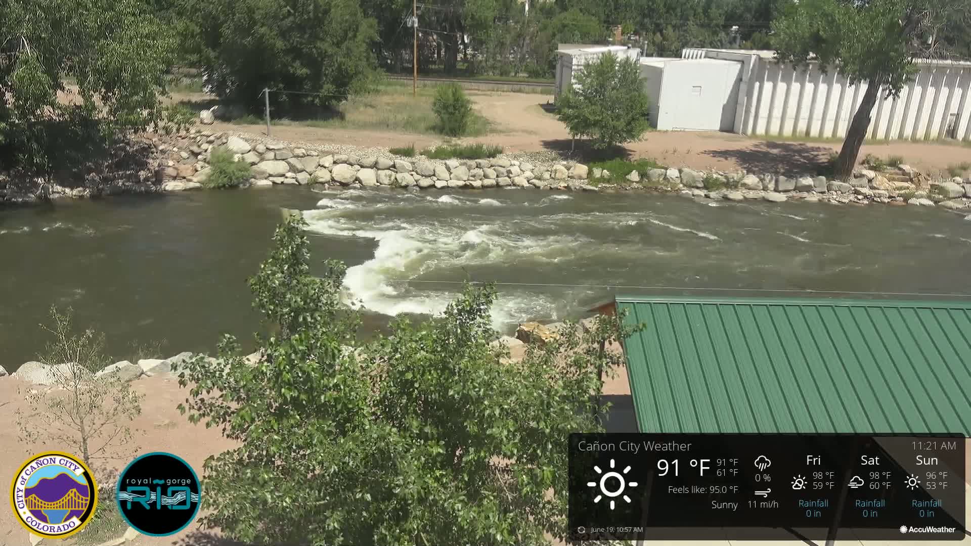 A swiftly flowing river, partially obstructed by a rock wall, is visible under sunny conditions with a green-roofed structure partially in view.