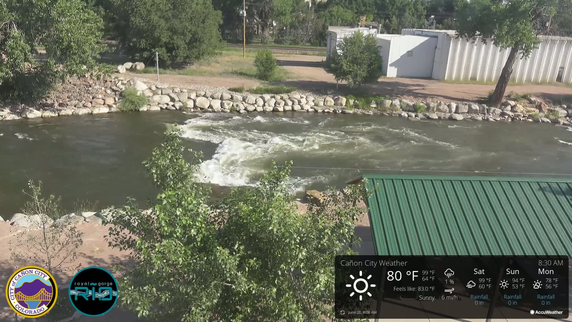 A sunny day shows a rocky riverbank with a swiftly flowing river and a green metal roofed structure partially visible in the foreground.