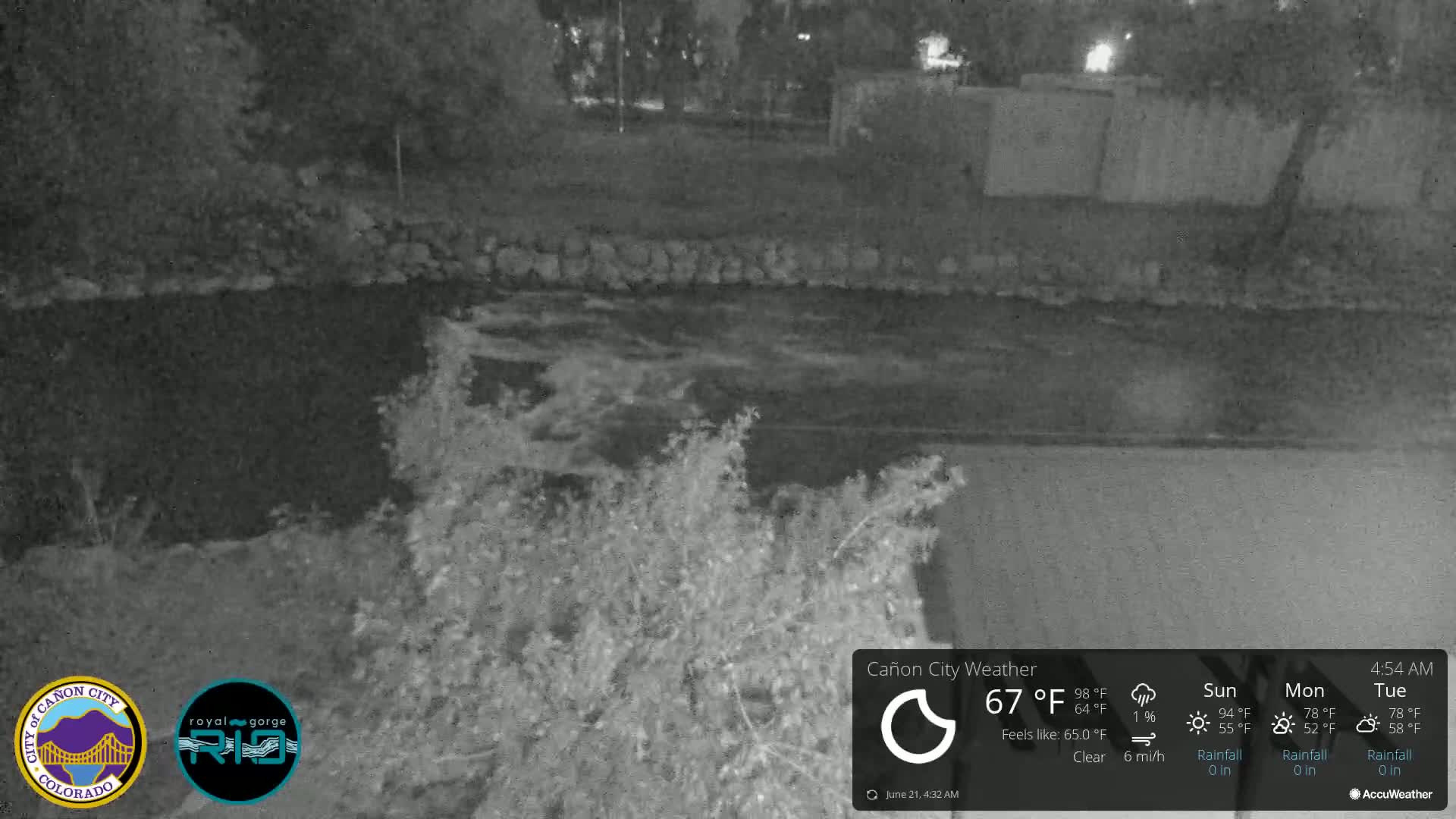 A nighttime, low-light view of a dark, slow-moving river bordered by vegetation and a low stone wall under clear skies.