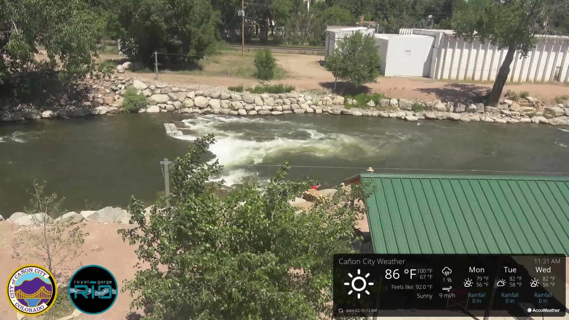 A sunny view of a river flowing over a low dam, with a green-roofed structure and lush green trees nearby.
