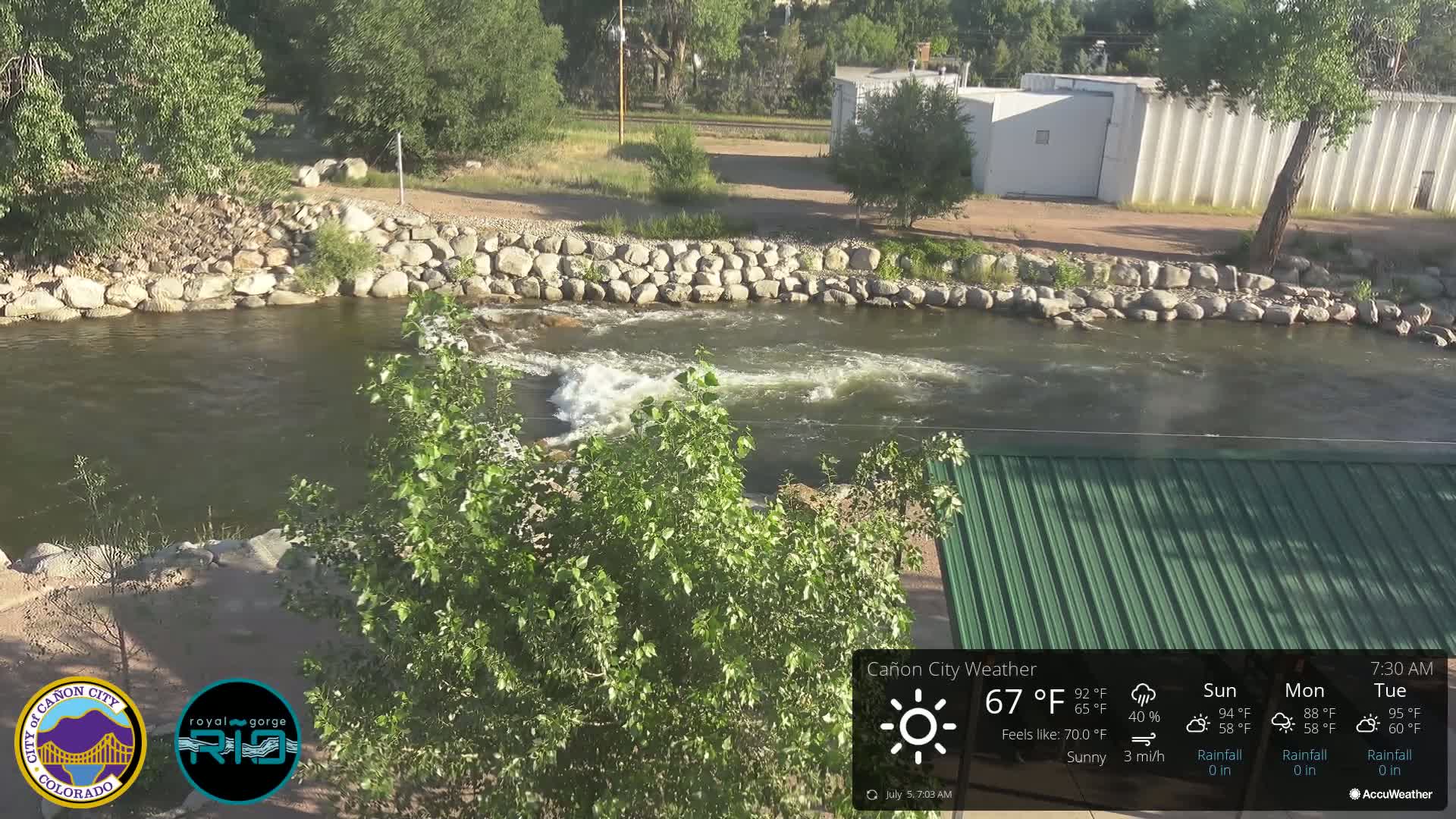 A sunny view of a river flowing past a rocky bank, with green vegetation and a partially visible green roofed structure.