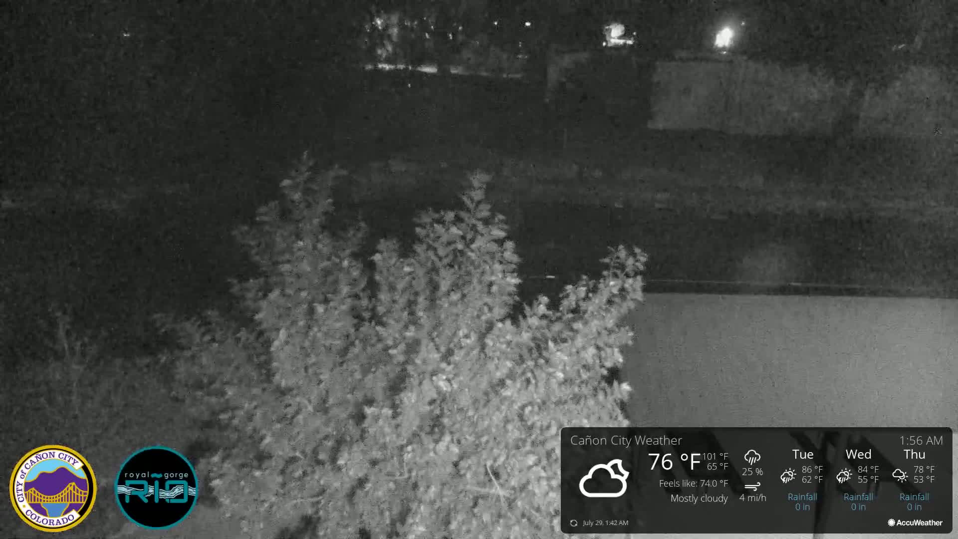 A nighttime, mostly cloudy scene shows dark shrubs in the foreground with a dark body of water and more dark land in the background.