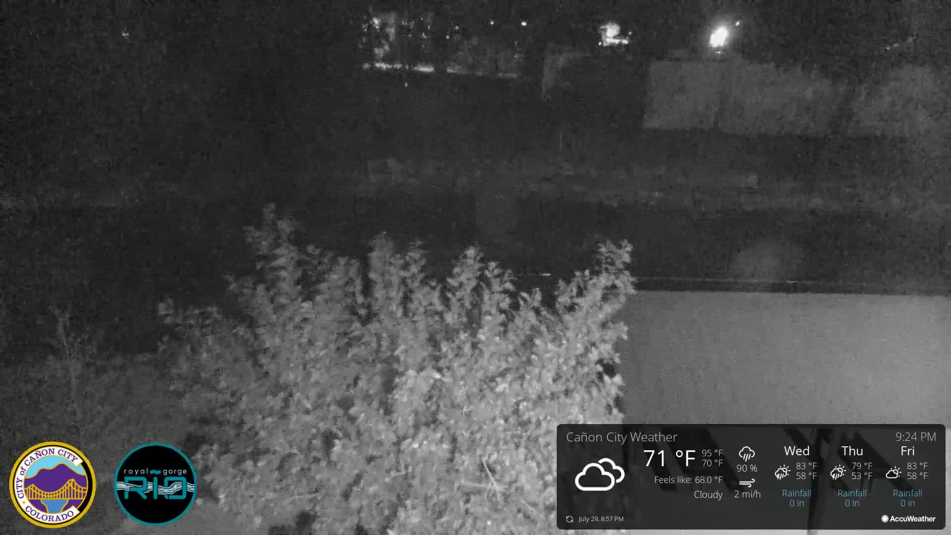 A nighttime, mostly cloudy scene shows dark shrubs in the foreground with a dark body of water and more dark land in the background.