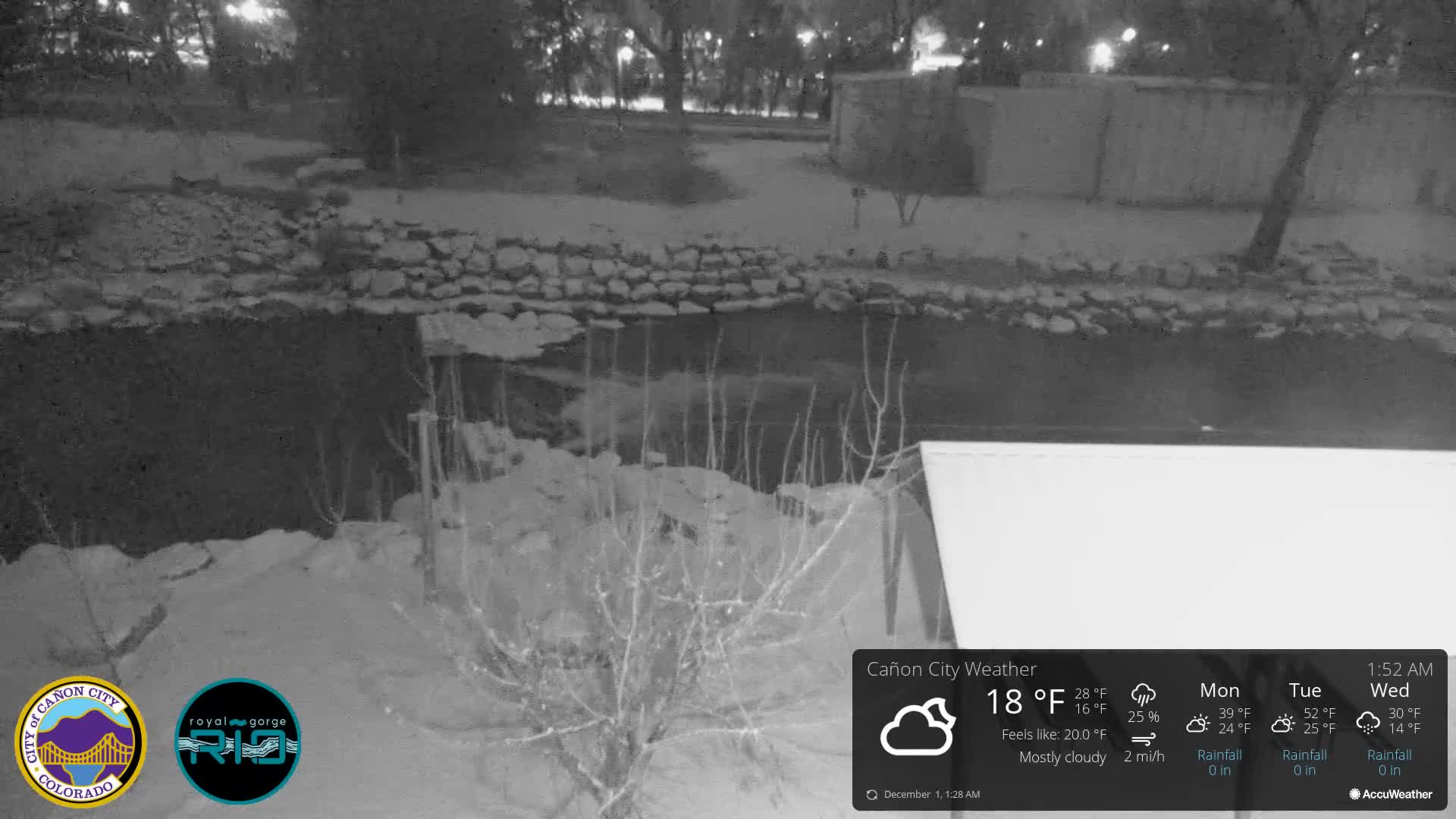 A black and white image captures a cold, cloudy winter night scene, showing a body of water with snow-dusted bare branches in the foreground, a rocky bank, and illuminated trees and structures in the background.