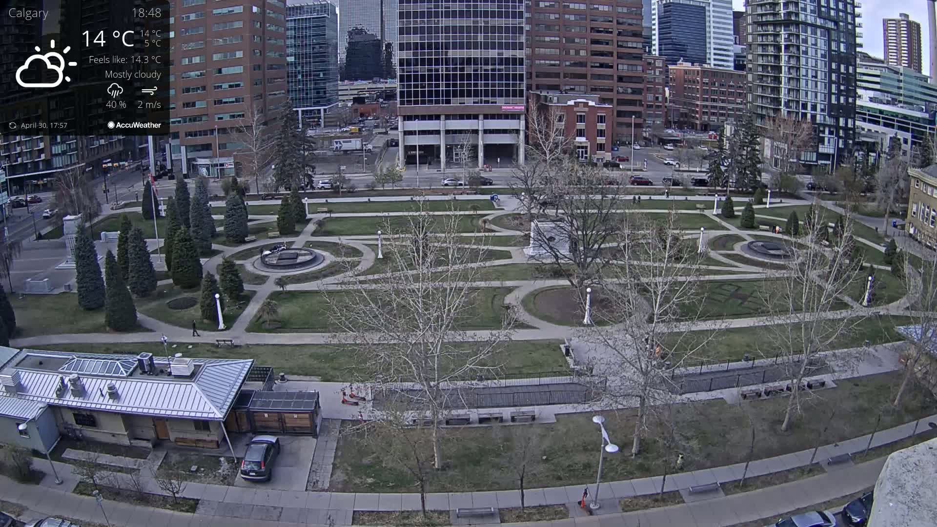 A mostly cloudy city park with a geometrically designed lawn, surrounded by buildings and bare trees.