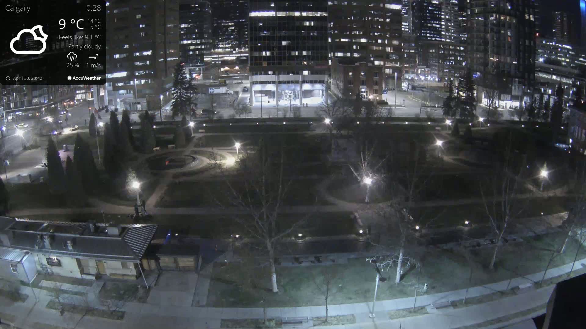 A nighttime, partly cloudy view of a city park with walkways, trees, and buildings in the background.
