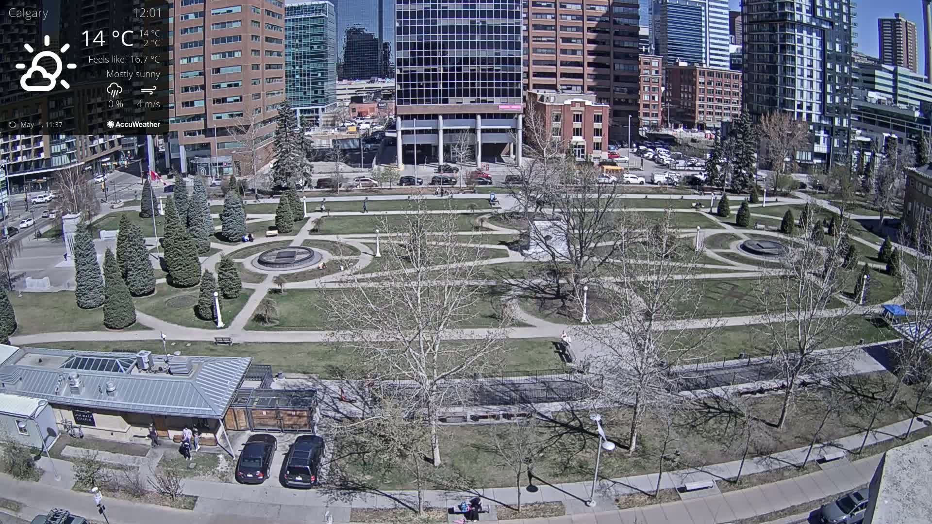 A mostly sunny city park with a geometrically designed lawn, bare trees, and several buildings in the background.