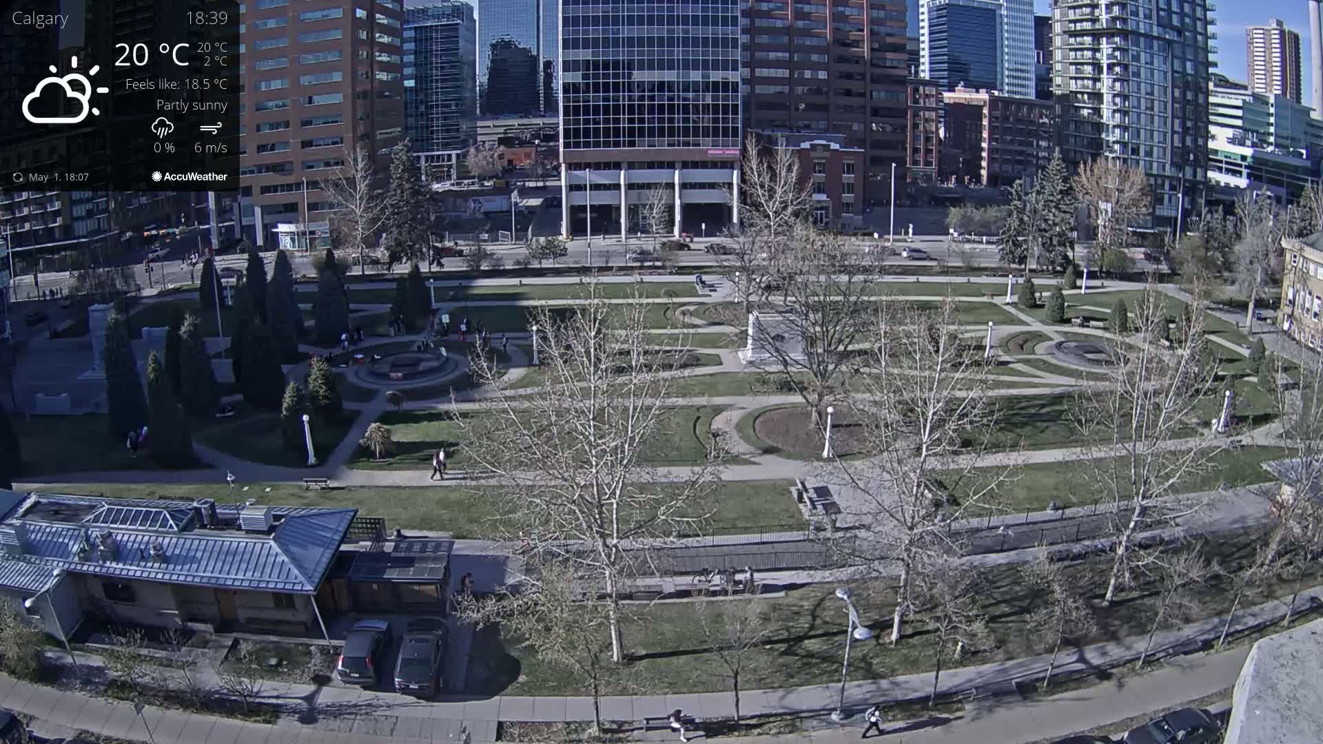 A partly sunny view of a city park with barren trees and a few people walking on paths, surrounded by tall buildings.