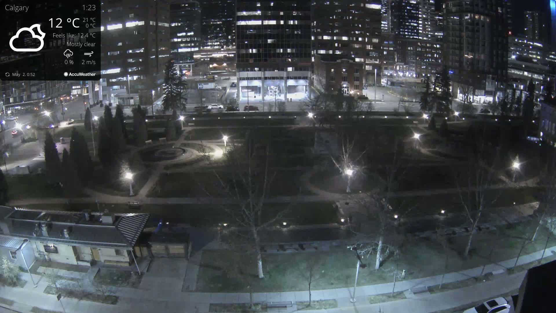A mostly clear night scene shows a park with lights illuminating pathways and trees, surrounded by city buildings.