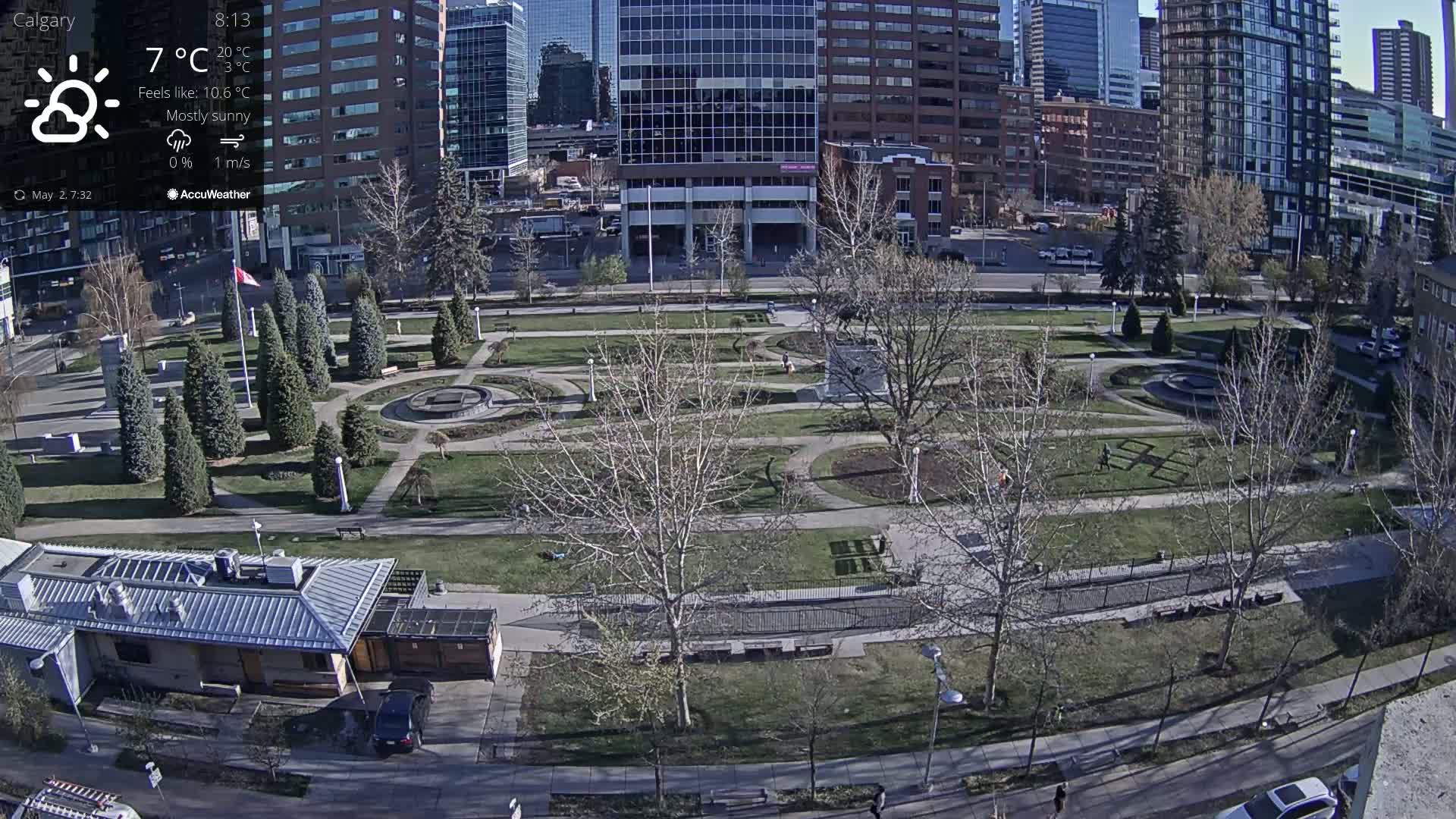 A mostly sunny city park with paved pathways, landscaping, and several buildings in the background.