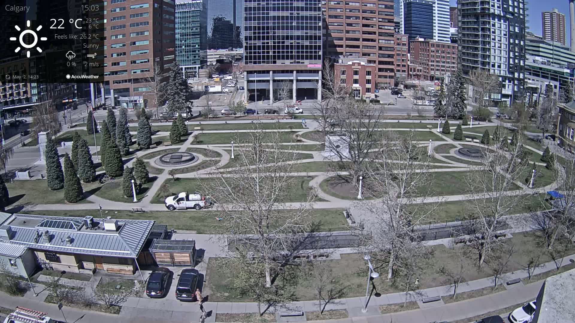 A sunny city park with a geometric layout of pathways and trees, surrounded by buildings, under a clear sky.