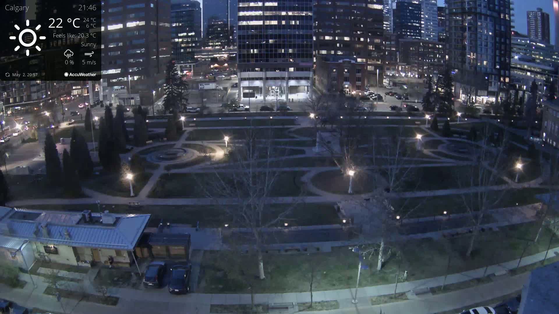 A sunny evening view of a city park with landscaped gardens, surrounded by city buildings and streets with cars parked along the sides.