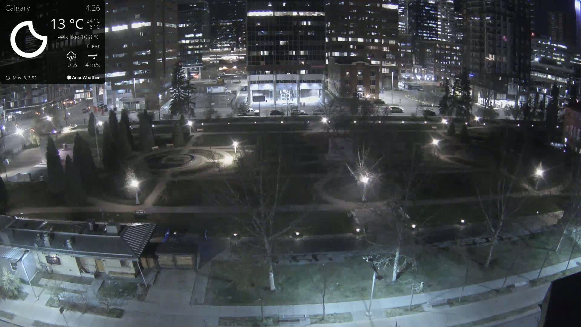 A nighttime view of a park illuminated by lamps, surrounded by city buildings under clear skies.