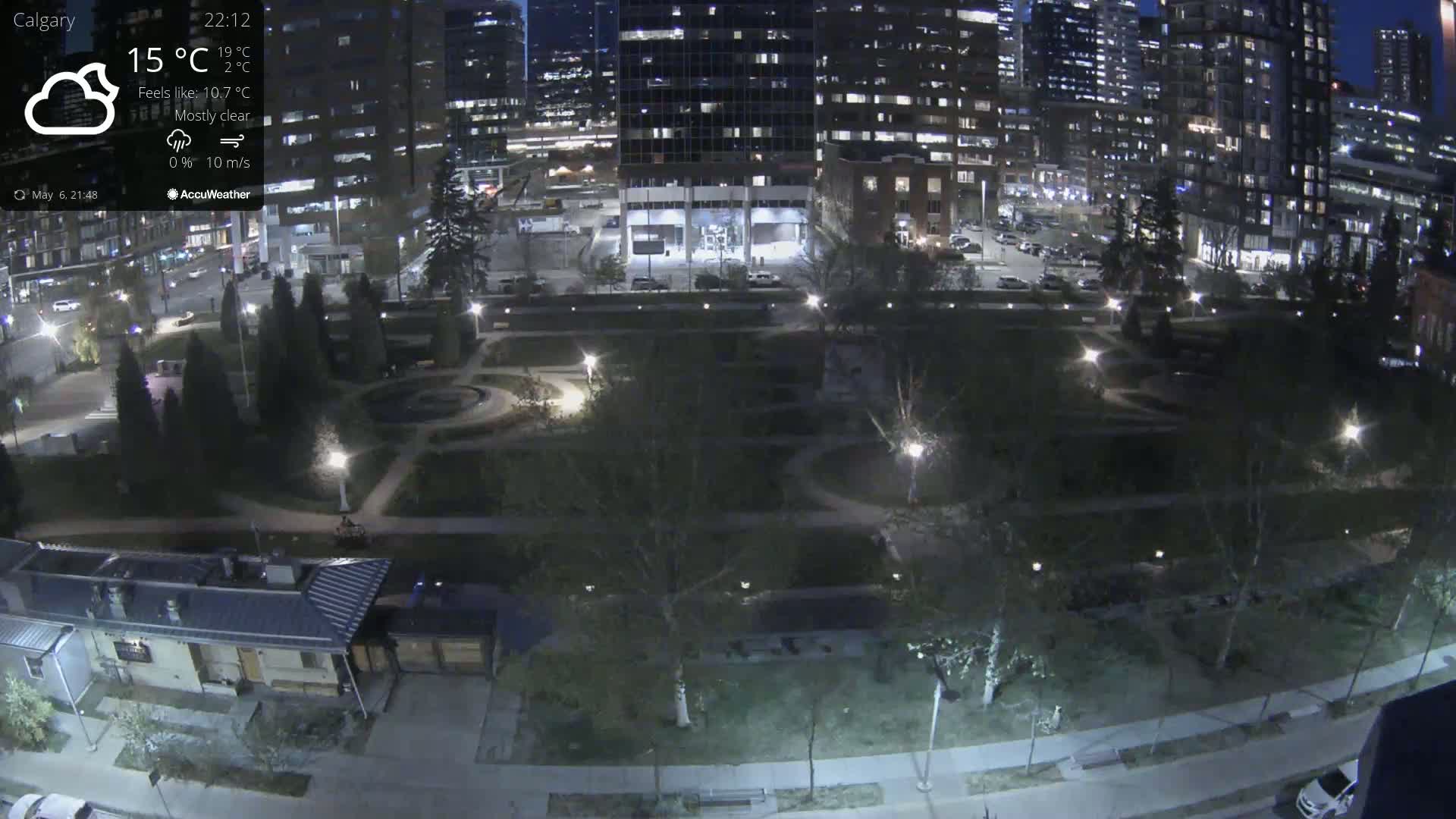 A nighttime aerial view of a city park, mostly clear with a 10 m/s wind, showing illuminated pathways, trees, and buildings in the background.