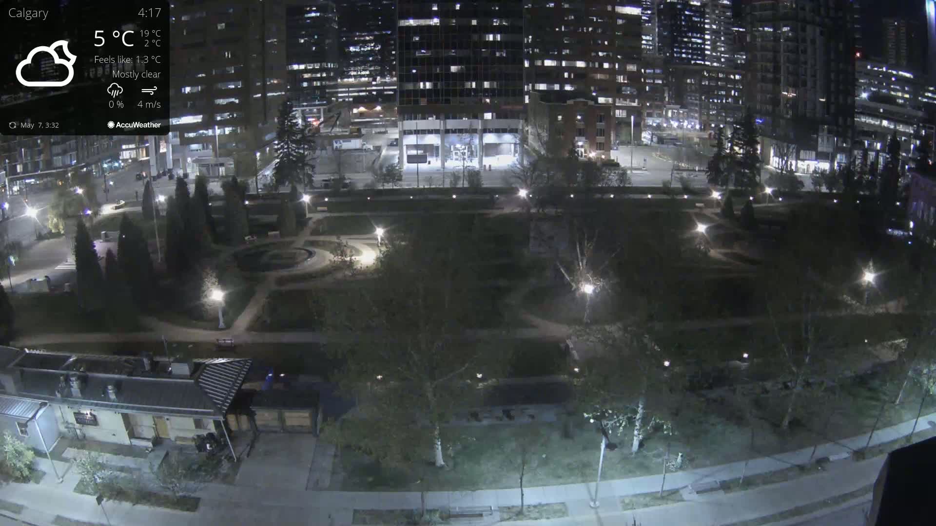 A mostly clear night scene shows a city park illuminated by lights, surrounded by tall buildings.