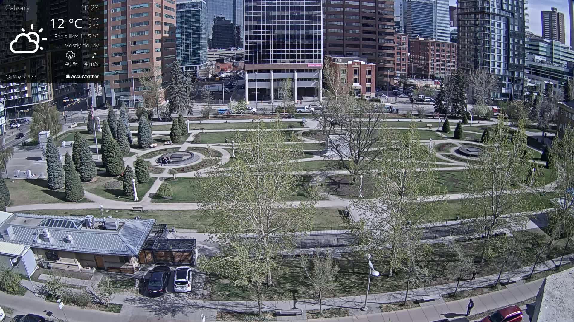 A mostly cloudy day reveals a city park with winding paths, young trees, and a small building, all surrounded by tall buildings.