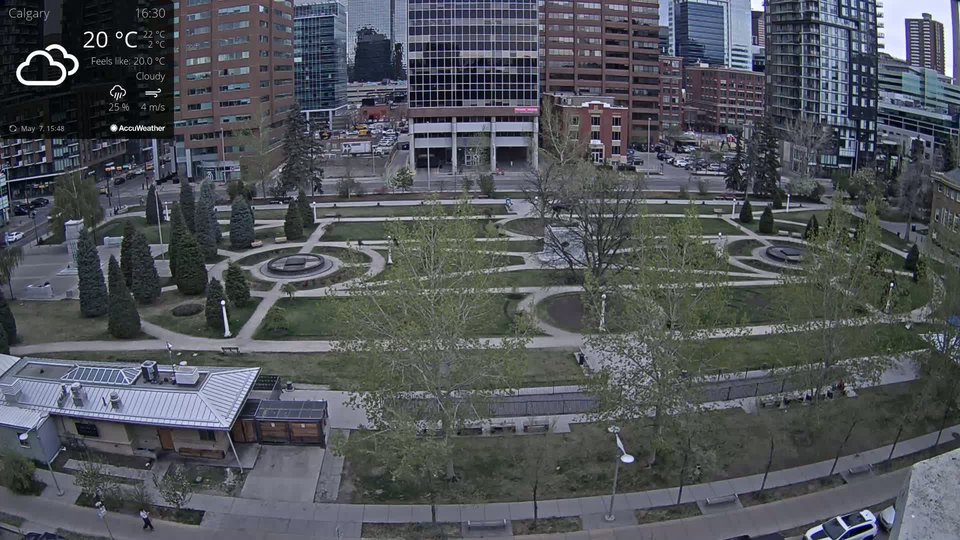 A city park with a geometric design, featuring paved pathways, grassy areas, and young trees, is situated under a cloudy sky.