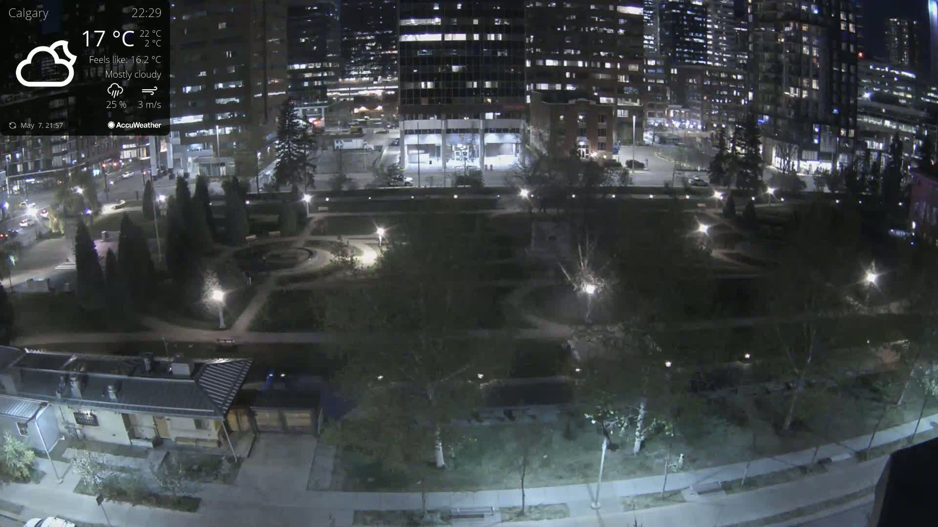 A mostly cloudy night scene shows a city park with pathways, landscaping, and lights, bordered by buildings and streets with some traffic.