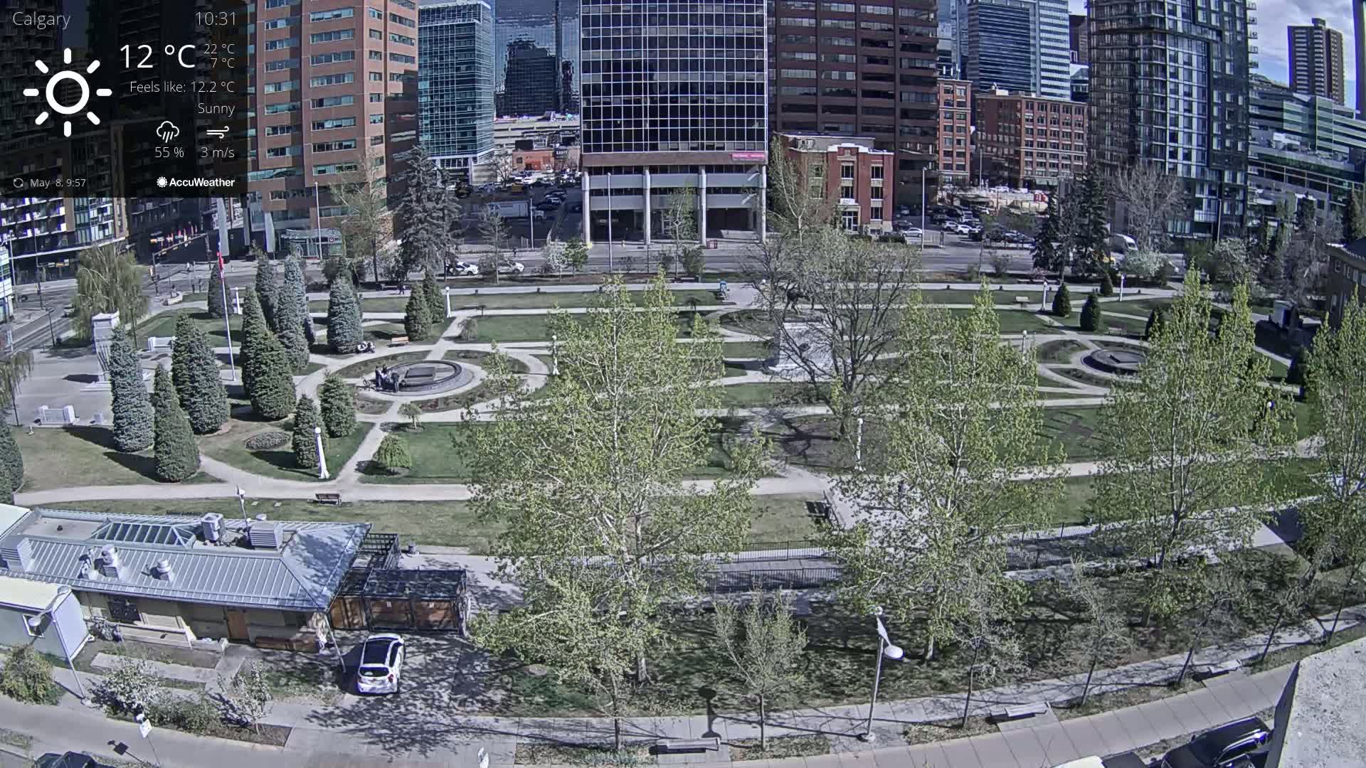 A sunny city park with paved pathways and young, leafy trees is situated between tall buildings under a partly cloudy sky.