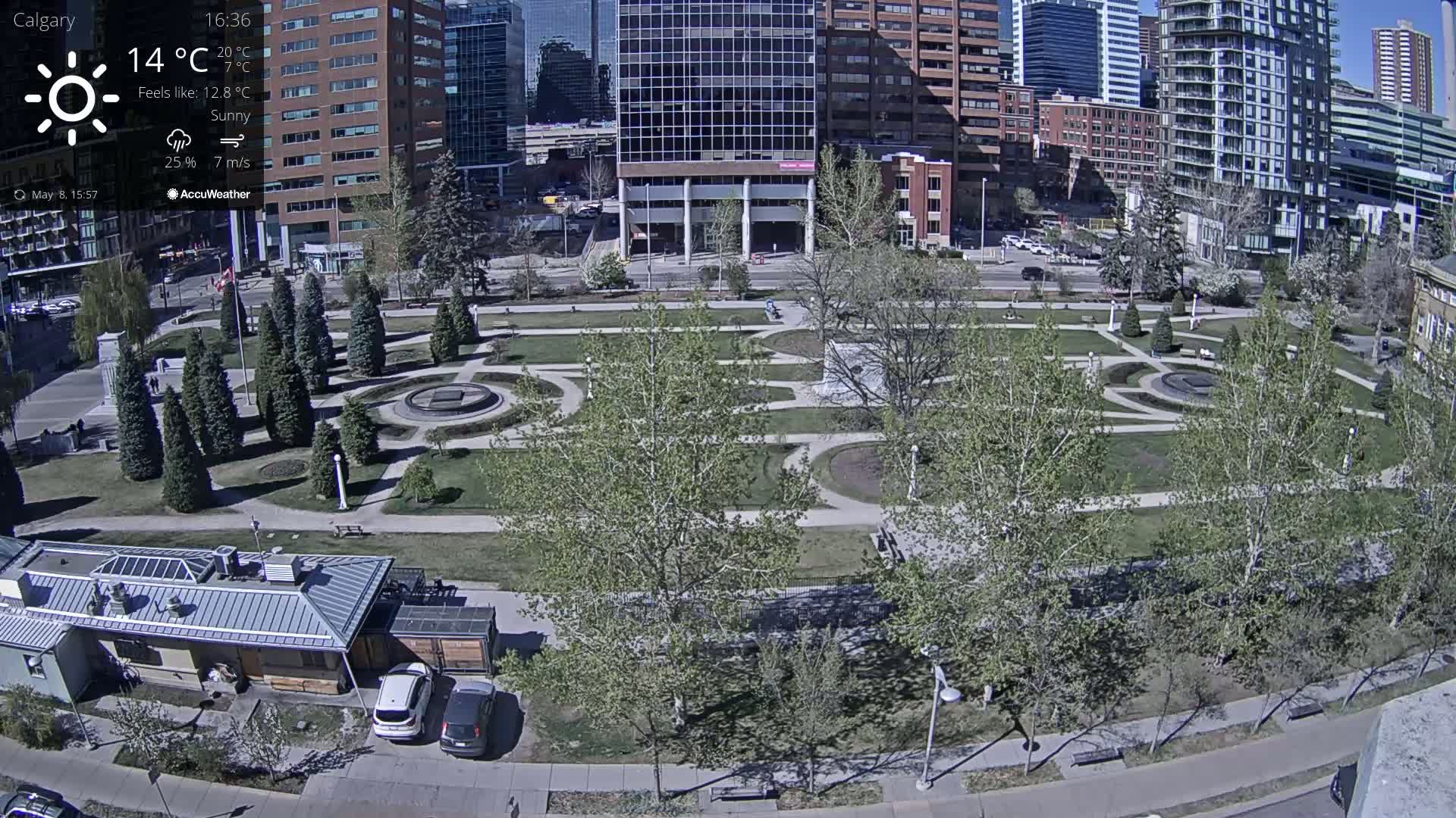 A sunny cityscape shows a park with paved pathways winding through grassy areas and young trees, surrounded by tall buildings.